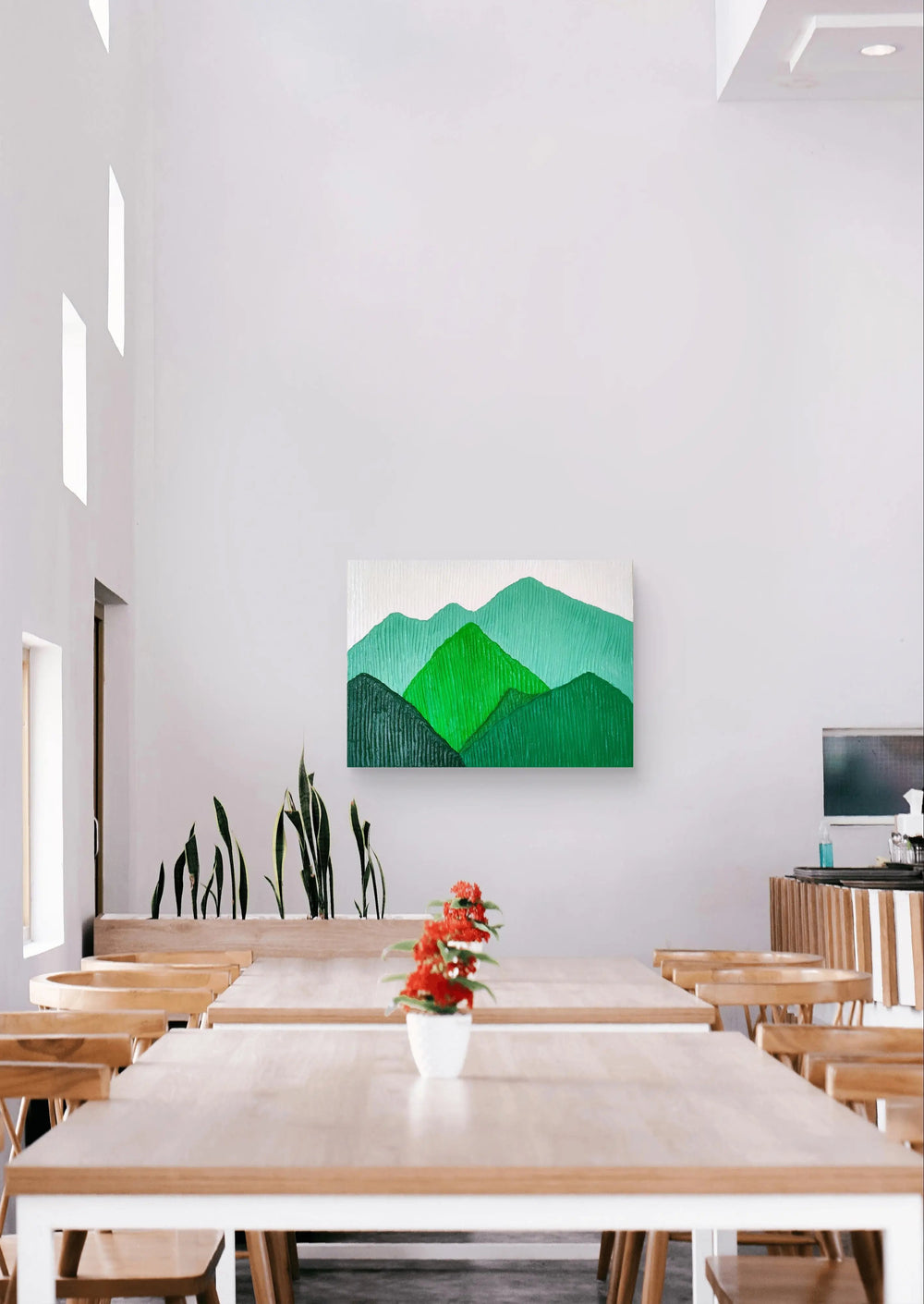 Green mountain landscape painting displayed in a bright cafe with wooden furniture.